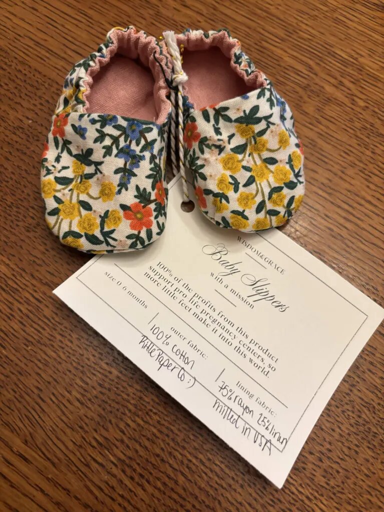 Pro-Life Baby Slippers in a cream wildflower print with pink linen lining, tied to a Wisdom & Grace tag